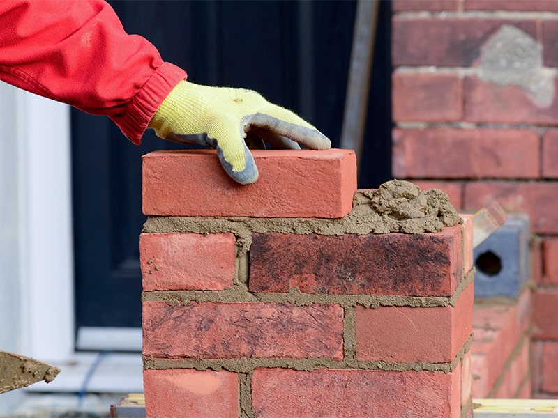 A gloved hand places a red brick atop a neat stack, with fresh mortar visible.