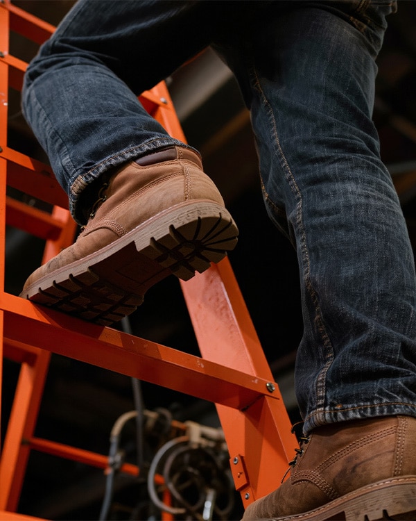 Close-up of a person wearing jeans and brown boots climbing a bright orange ladder.